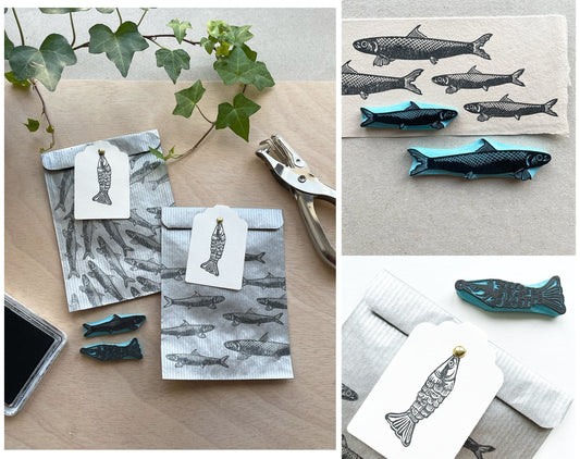 Hand carved sardine rubber stamps in three sizes used to decorate kraft envelopes and gift bags with a repeated fish pattern, ink pad and white gift tags on a wooden table